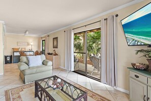 Ein Wohnzimmer mit Sofa, Couchtisch und Fernseher. Eine Glastür öffnet sich zu einem Balkon mit Gartenmöbeln.