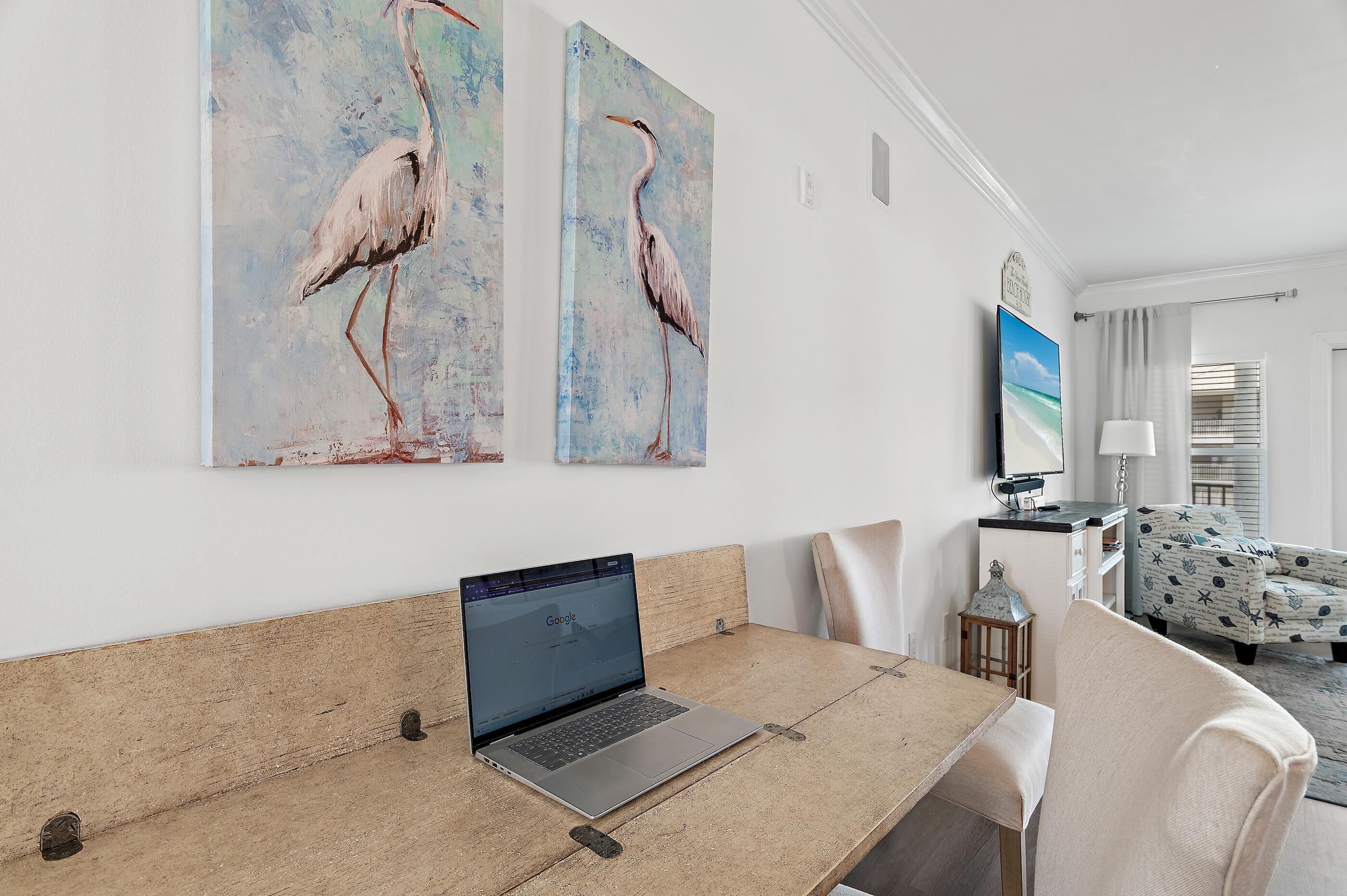 A room with a laptop on a table, two paintings of birds, a TV, and a patterned sofa.