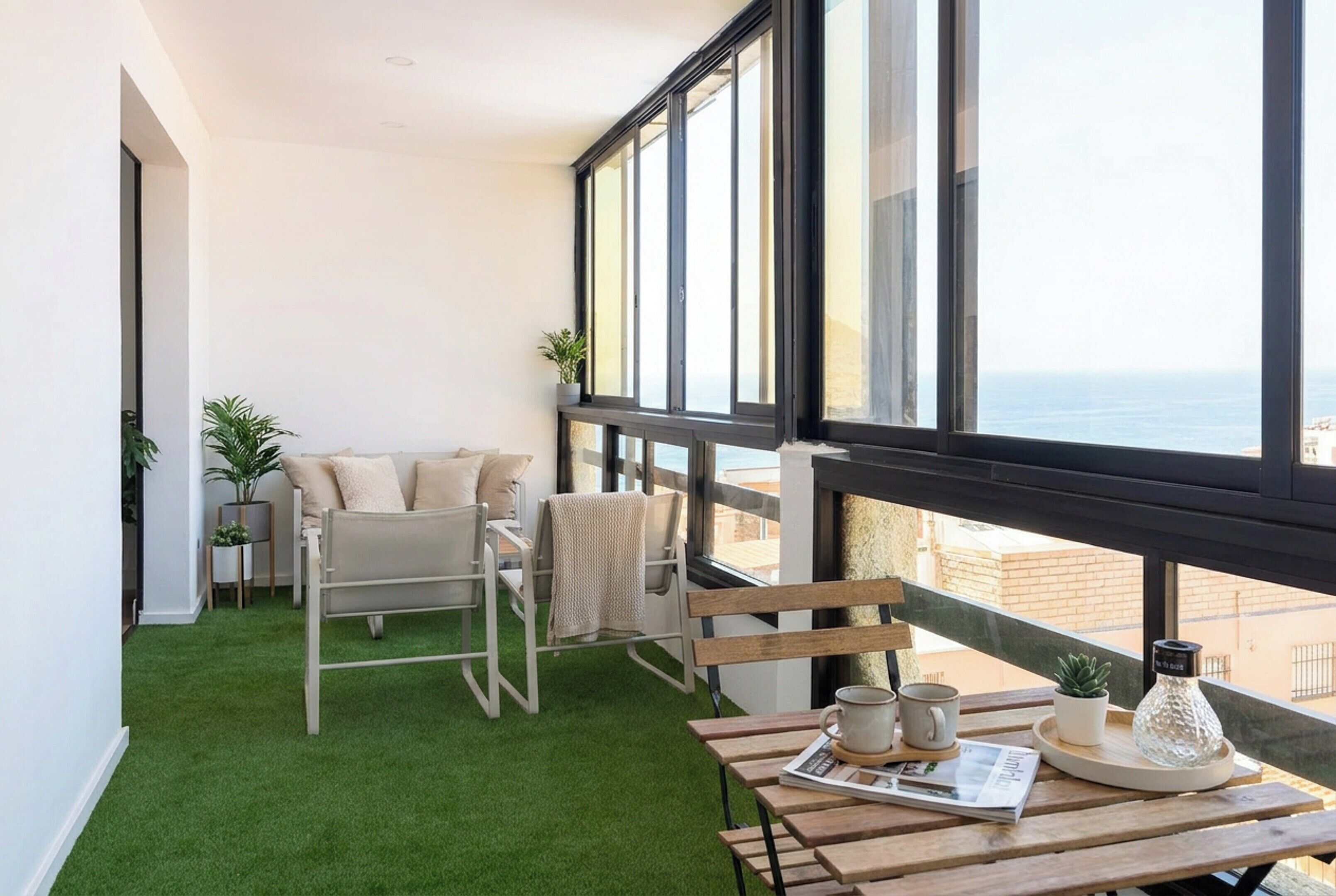 A balcony with a view of the sea, a sofa, a table with a newspaper, and two potted plants.