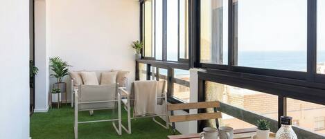 A balcony with a view of the sea, a sofa, a table with a newspaper, and two potted plants.