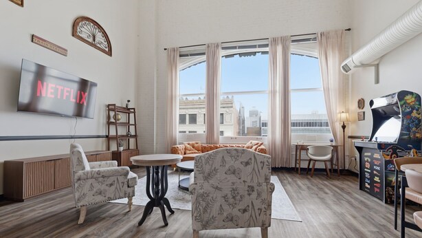 A modern living room with a flat-screen TV, a sofa, a gaming table, and a view of buildings through large windows.
