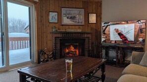 A cozy room with a fireplace, wooden walls, and a large window overlooking a snowy deck. - Family friendly 3 bedroom condo minutes from Mount Snow. (Dover)
