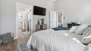 Une chambre avec un grand lit, un téléviseur fixé au mur, une table de chevet avec une lampe et une porte donnant sur une salle de bain.
