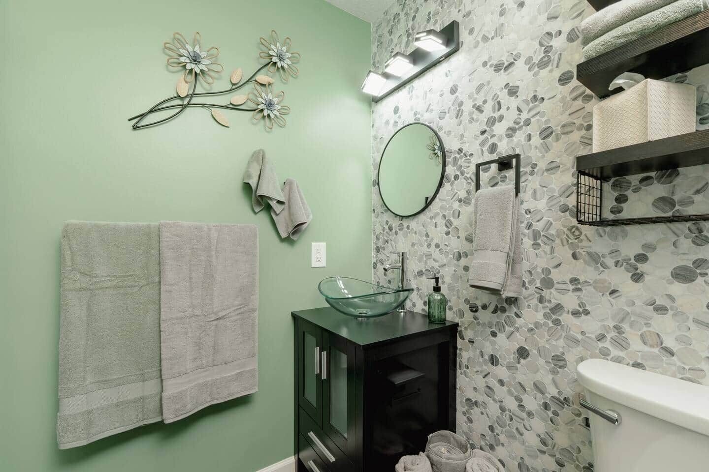 A bathroom with green walls, a glass bowl sink, a black cabinet, two towels, a mirror, and a towel rack.