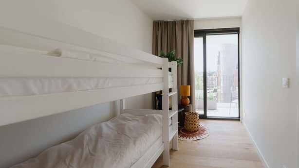 Une chambre avec un lit superposé, une petite table avec une lampe, une plante et une vue sur la ville par une grande fenêtre.
