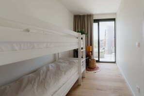 Quarto com beliche, uma mesinha com abajur, uma planta e vista da cidade por uma janela grande.