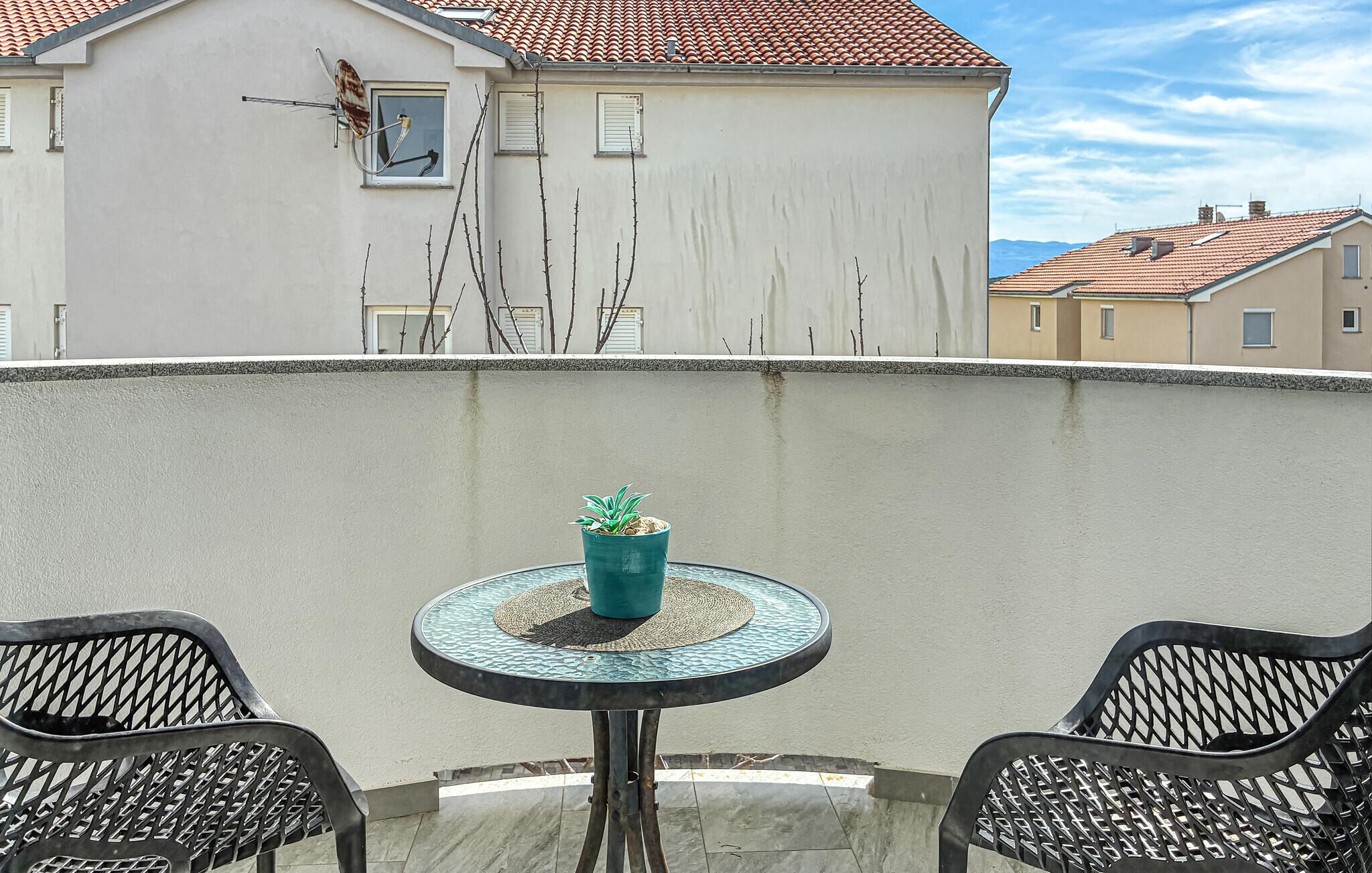 Een balkon met een ronde tafel, twee stoelen, een bloempot en een gebouw met een pannendak op de achtergrond.