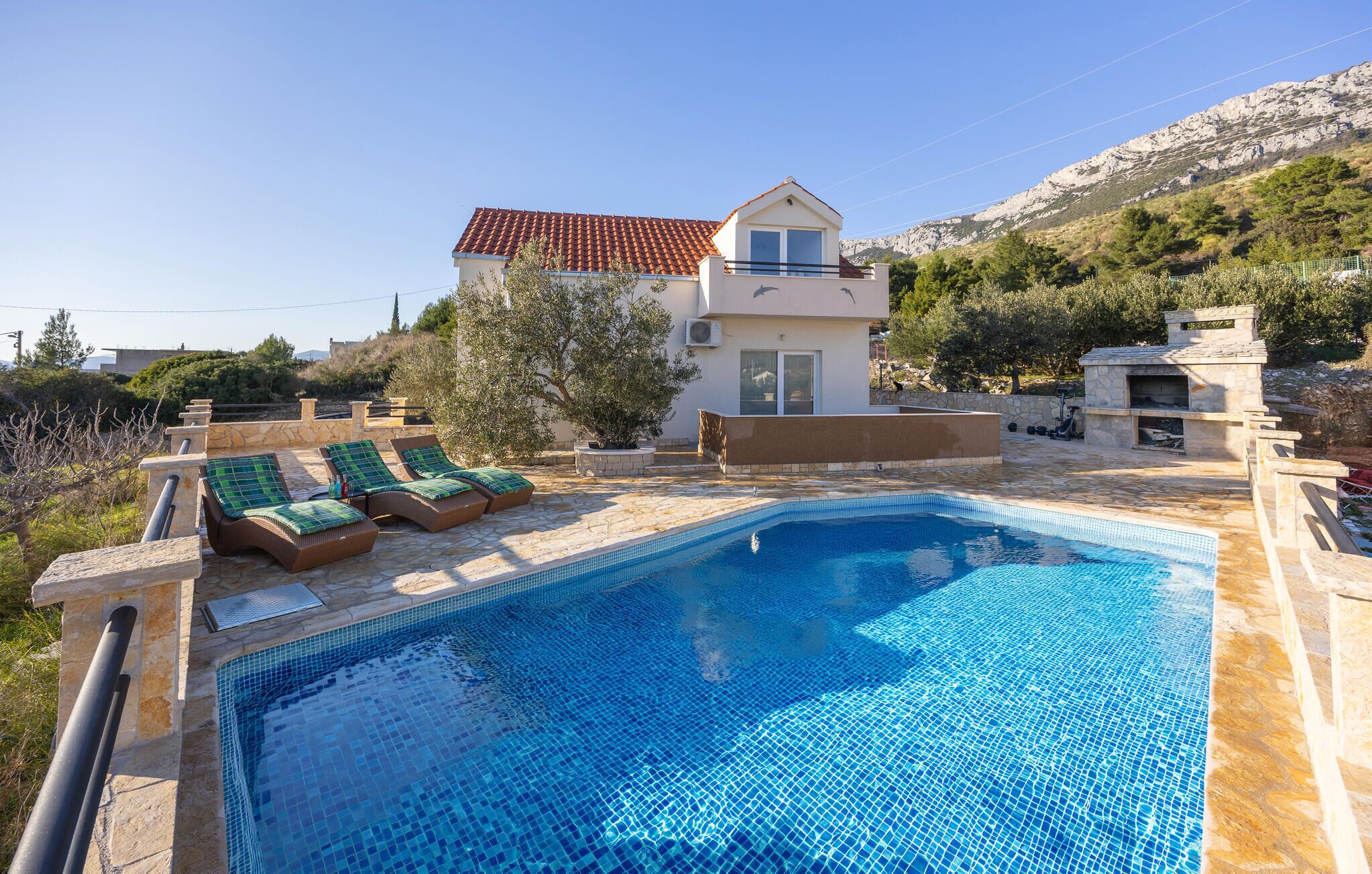 A swimming pool with lounge chairs, a stone deck, and a house with a tiled roof in the background.