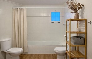 A bathroom with two toilets, a bathtub, a shower curtain, a window, and a wooden shelf with a basket. - 1 bedroom awesome apartment in Denia (Denia)