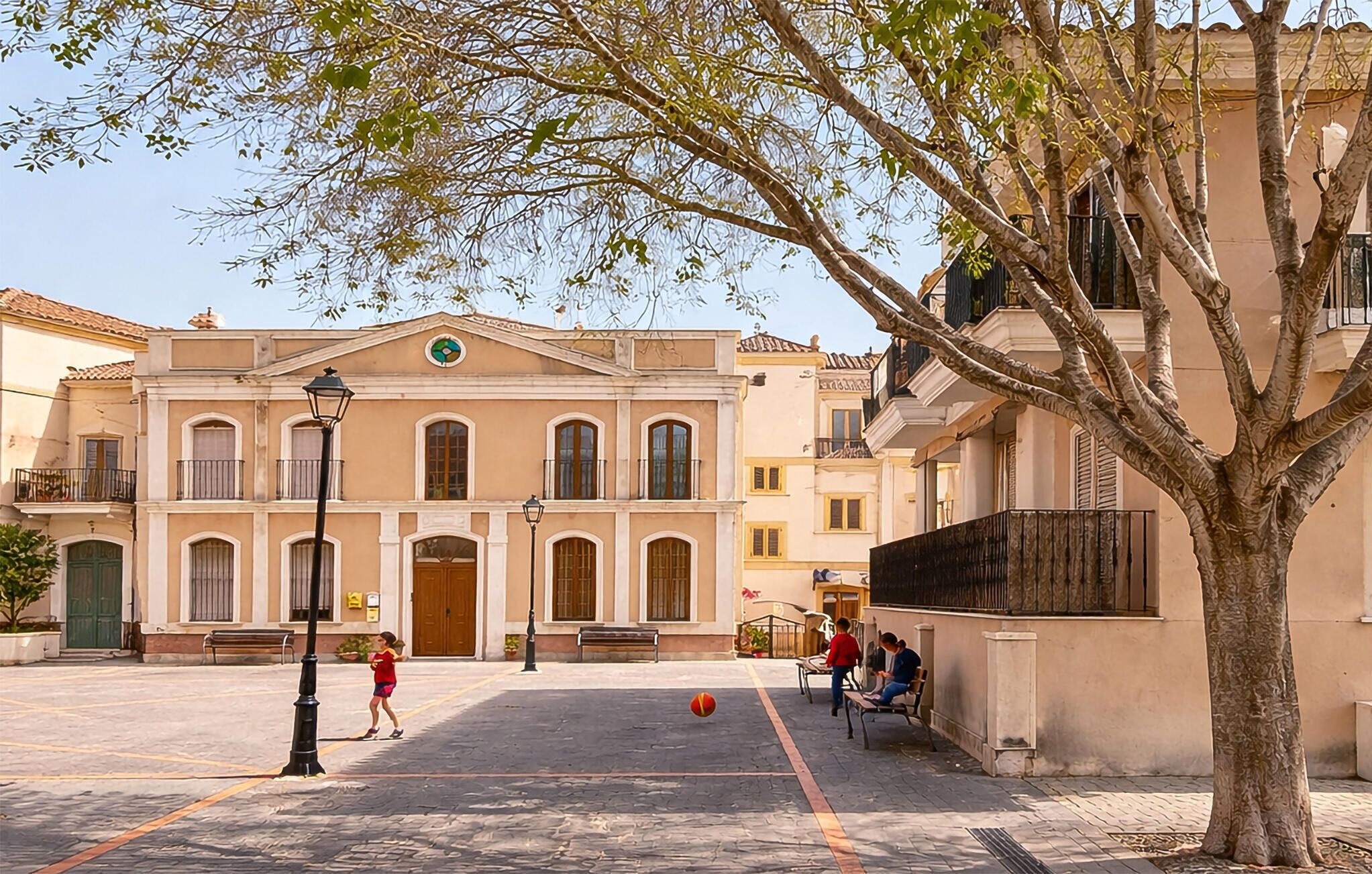 Un patio con un edificio que tiene una entrada principal, un árbol y una cancha de baloncesto.
