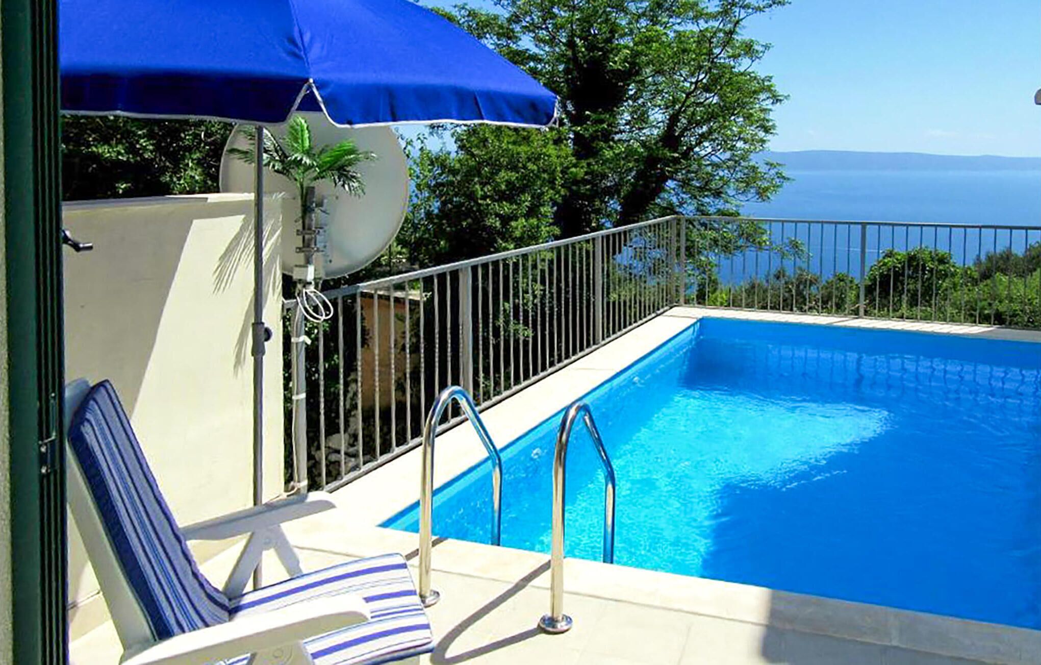 A swimming pool with a view of the sea, a lounge chair, and a blue umbrella.