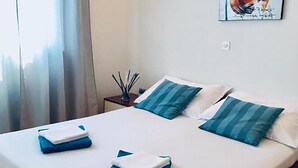 Ein ordentlich bezogenes Bett mit blau-weißer Bettwäsche, ein kleiner Nachttisch mit einer Lampe und ein Fenster mit Vorhängen.