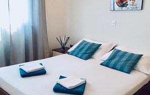 A neatly made bed with blue and white bedding, a small bedside table with a lamp, and a window with curtains. - Amazing home in Makarska (Makarska)