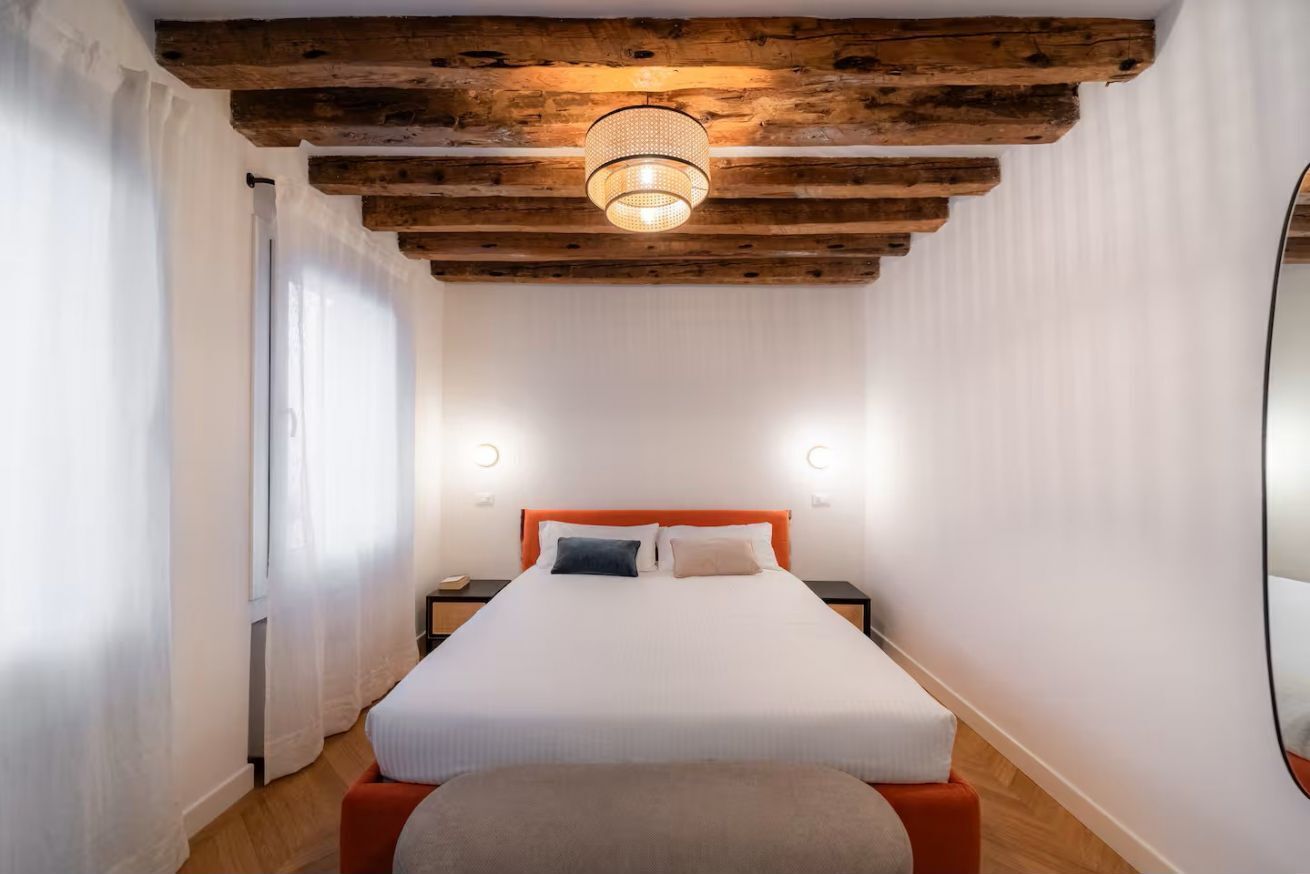 Une chambre avec un plafond en bois, un lit avec une literie blanche, deux tables de chevet et un grand miroir au mur.