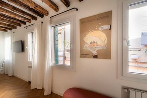 A room with wooden beams on the ceiling, a flat-screen TV mounted on the wall, a window with white curtains, and a poster of a building. - Cà del Bacino, San Marco (Venice)