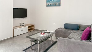 Ein modernes Wohnzimmer mit Flachbildfernseher, Glastisch, grauem Sofa und einem kleinen Beistelltisch mit Schale und Fernbedienung.