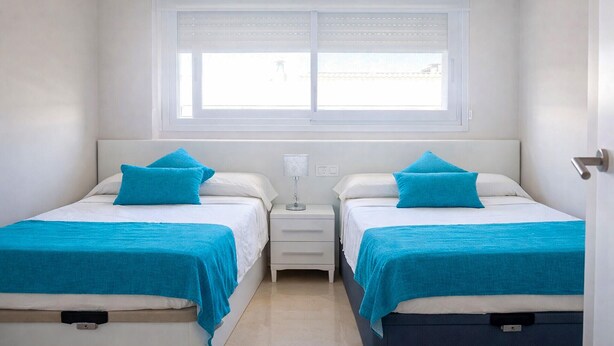 A room with two beds, each with a white mattress and teal bedding. There is a window with blinds, a bedside table with a lamp, and a door on the right. - 3 bedroom gorgeous apartment in Cabanes (Cabanes)