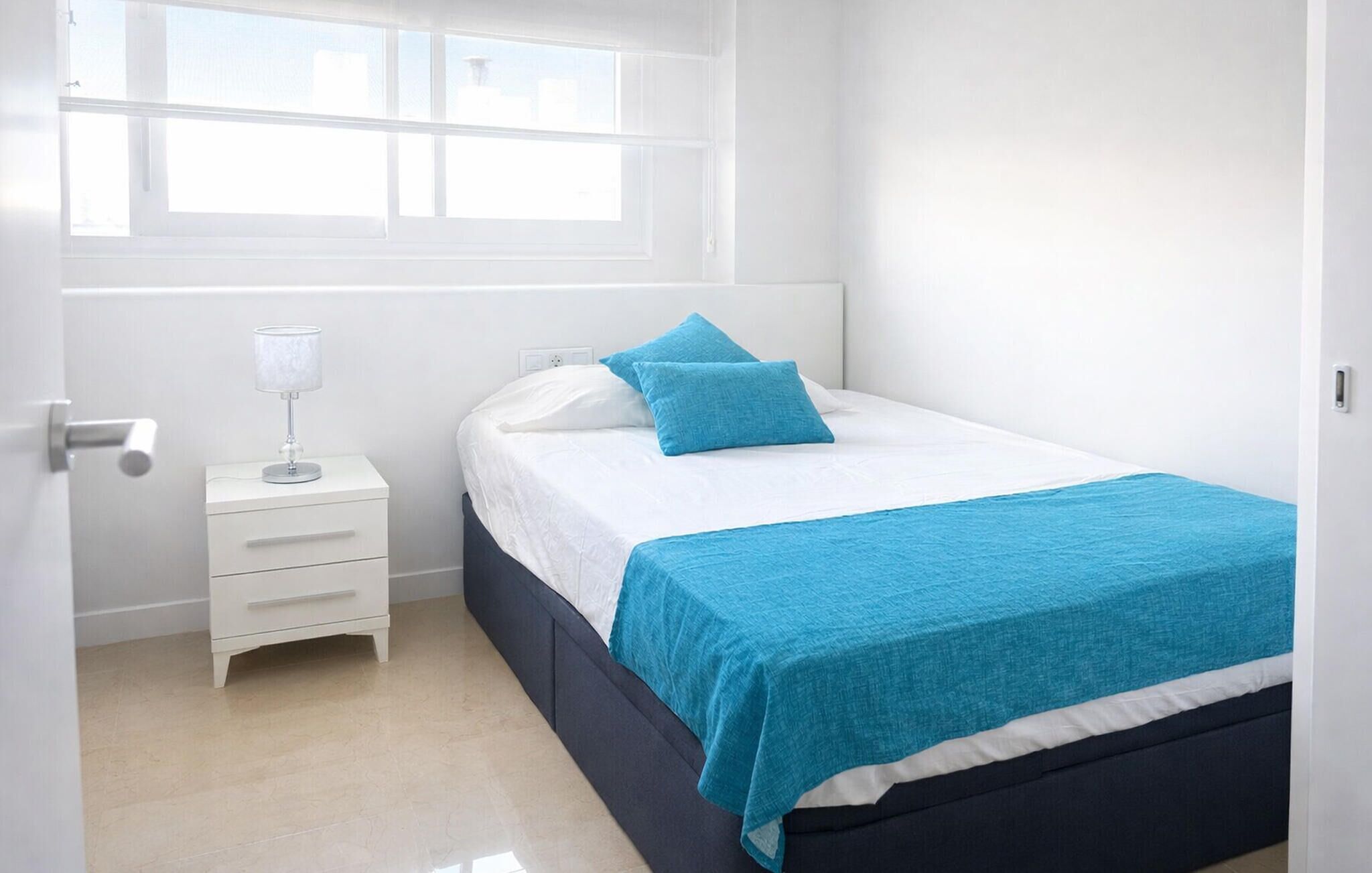 A bedroom with a bed, a bedside table with a lamp, and a window.