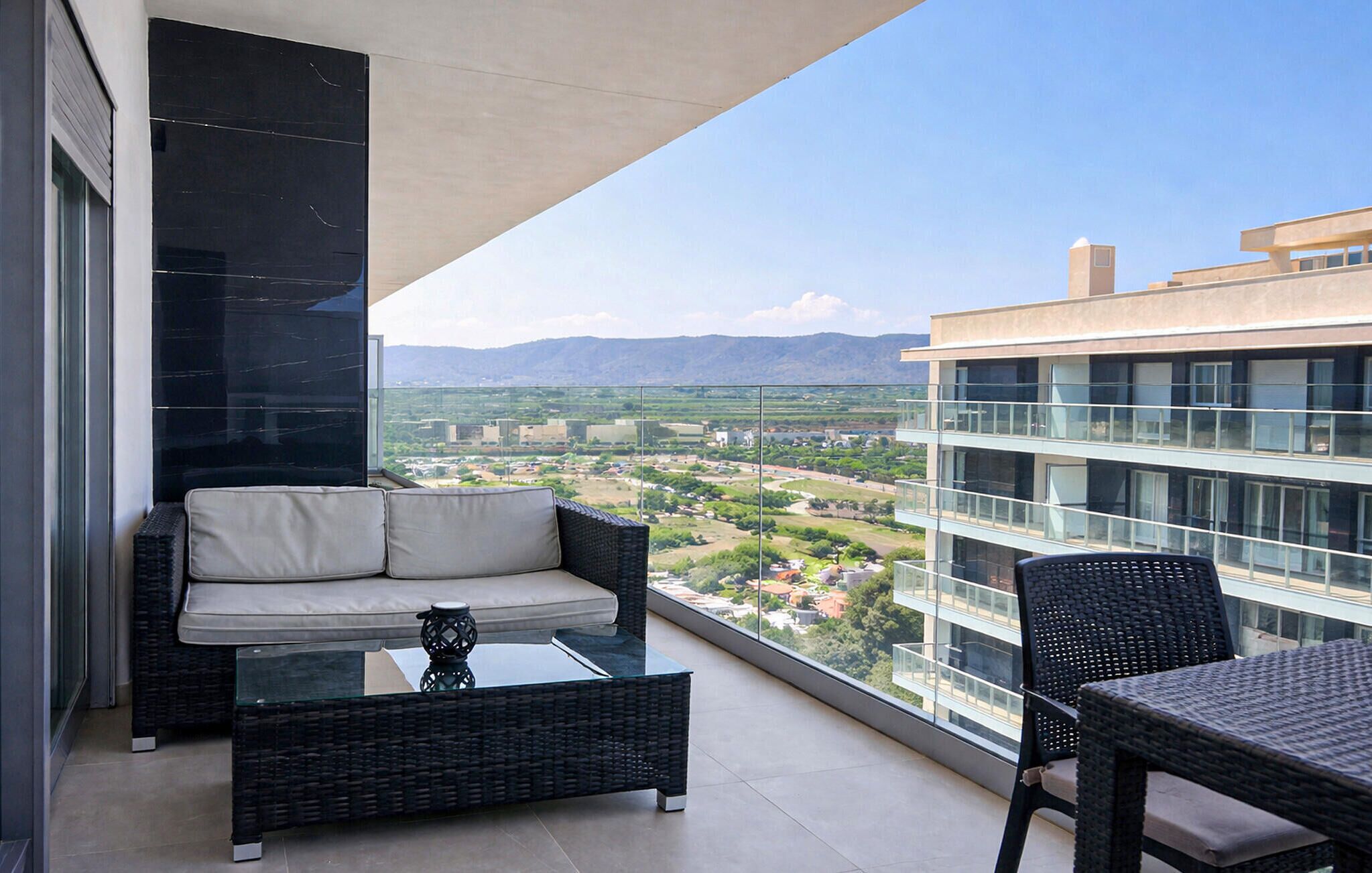 A balcony with a sofa, a small table, and a chair, overlooking a cityscape with buildings and mountains.