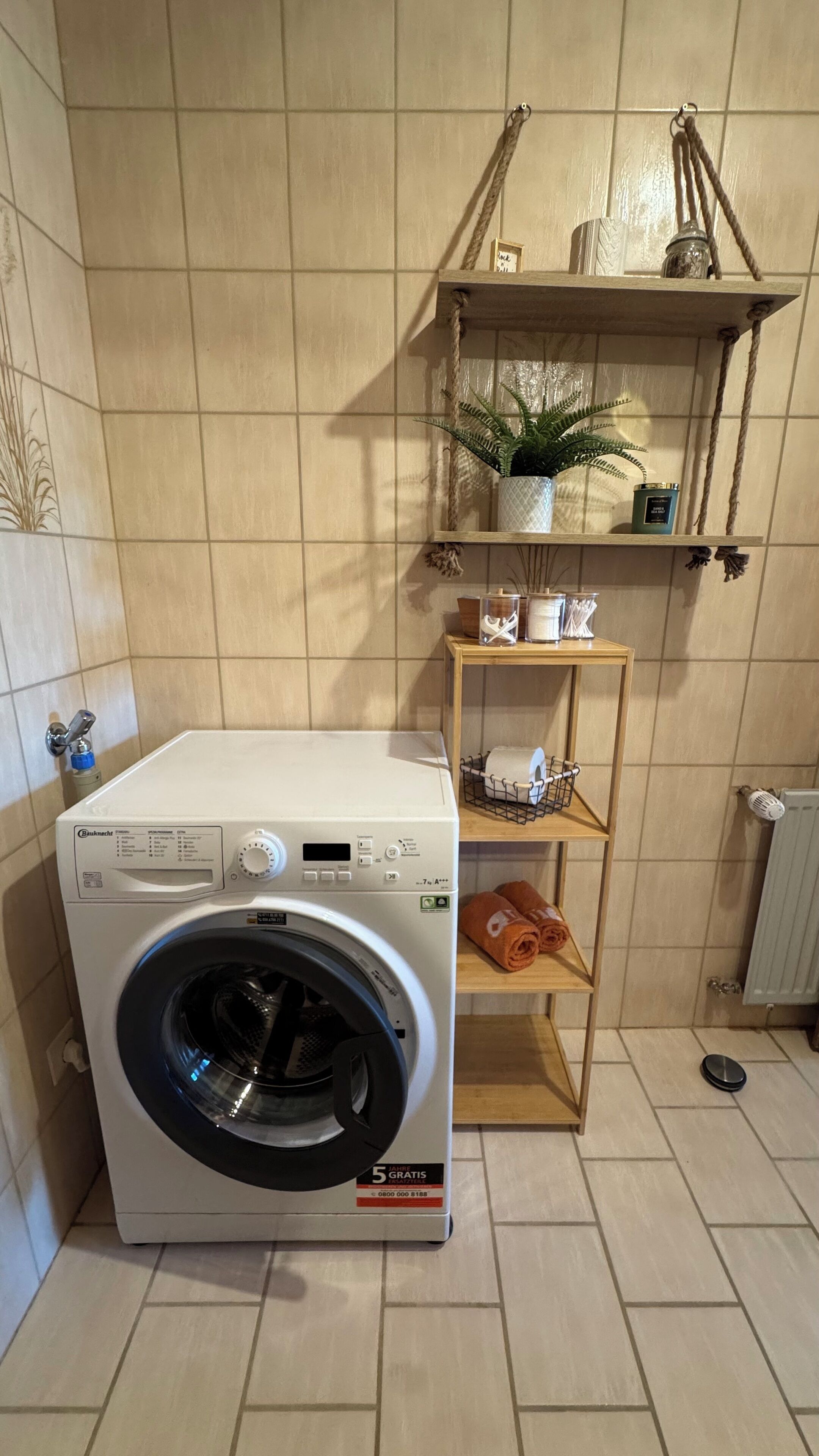 Een compacte, witte wasmachine met frontlader in een kleine wasruimte met betegelde muren, een houten plankje met een bloempot en een radiator.