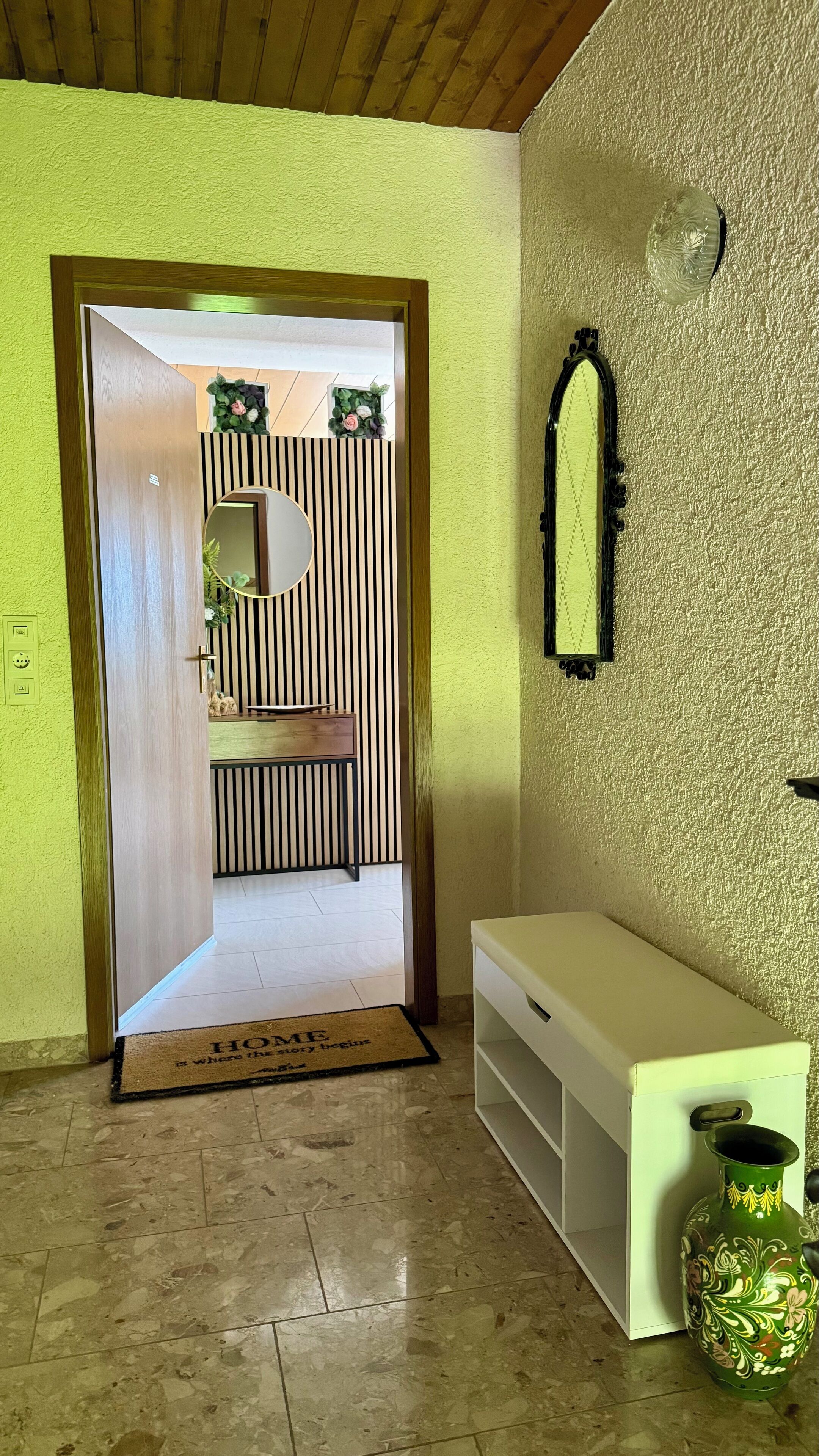 Um corredor com uma porta de madeira que dá para um quarto com parede listrada e um espelho. É possível ver uma mesa consola branca e um vaso verde.
