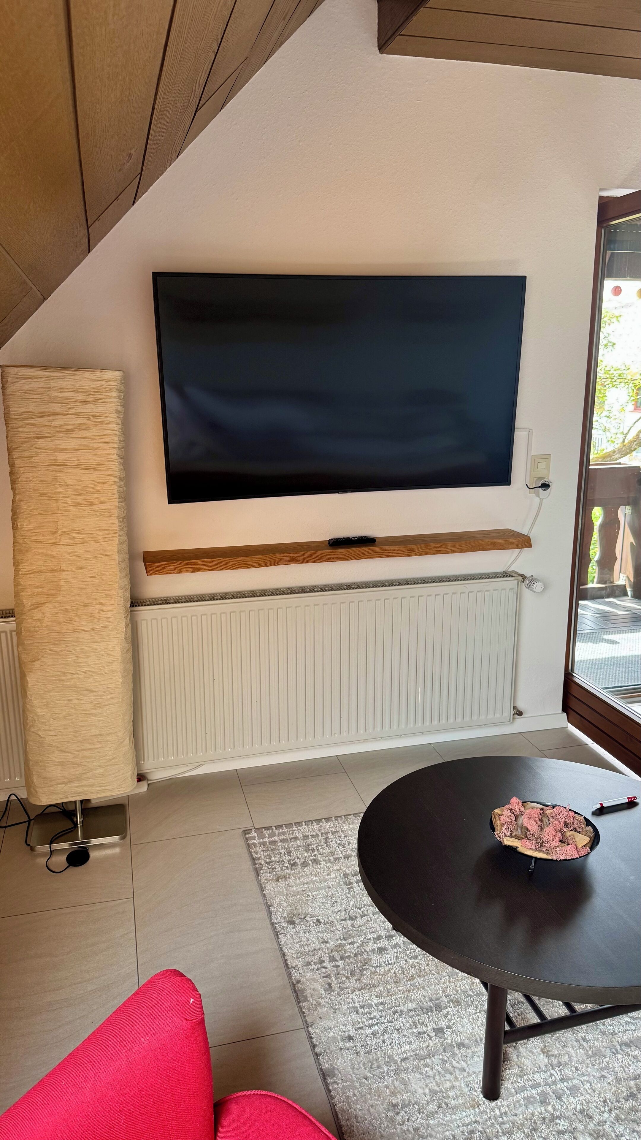 Een woonkamer met een grote flatscreen televisie aan de muur, een houten plank eronder, een radiator eronder en een ronde tafel met een schaal roze bloemen.