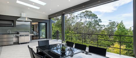 A modern kitchen with a large island, a dining area with a glass table, and a balcony with a view of trees.