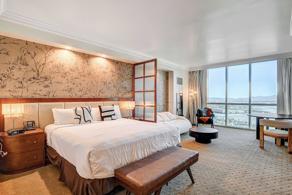 Hotelkamer met een groot bed, nachtkastjes, een bankje en uitzicht op de stad, met een groot raam.