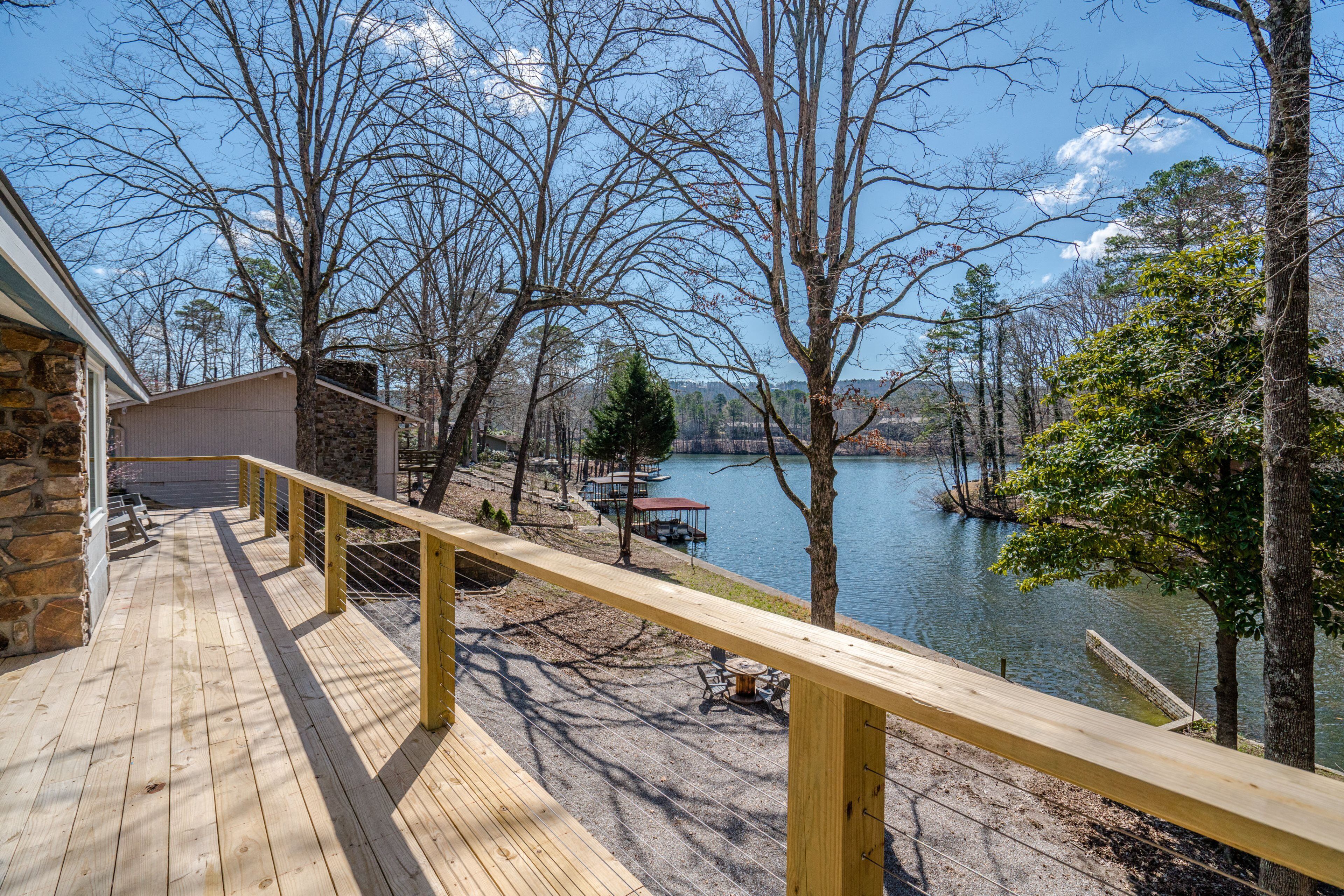 Une terrasse en bois avec une balustrade donnant sur un lac, entourée d’arbres et d’un bâtiment en pierre.