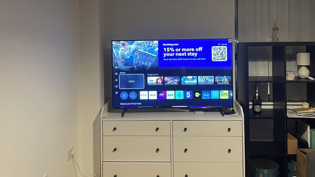 A modern living room with a flat-screen TV displaying a booking offer, a white cabinet with drawers, a wooden coffee table, and a pendant light. - City Centre Sunniside Sunderland (Sunderland)