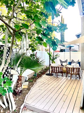 Une terrasse en bois, agréée de mobilier d’extérieur, entourée d’une végétation luxuriante et d’une clôture blanche.