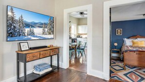 Un salon avec un grand téléviseur fixé au mur, une console en bois et une vue sur la cuisine.
