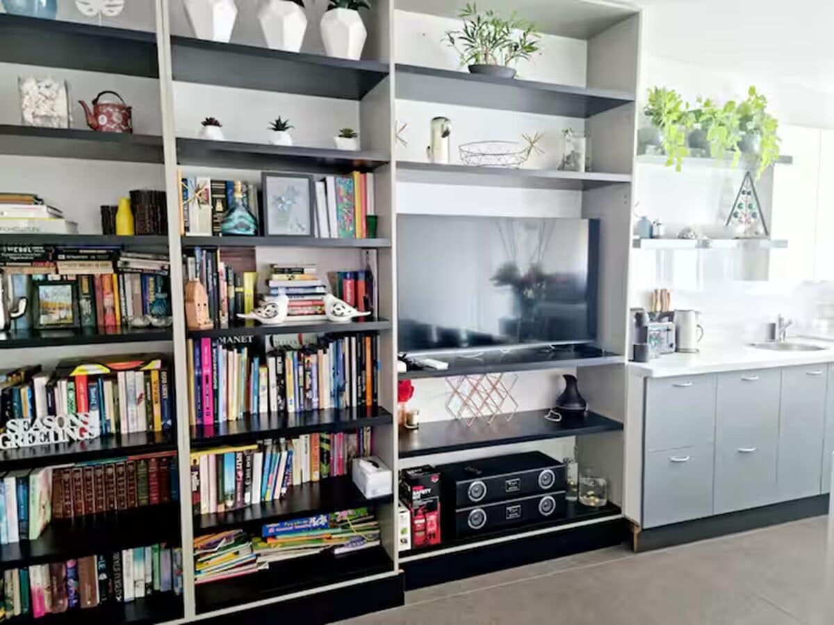 A modern living room with a large flat-screen TV, a bookshelf filled with books, and a kitchen area with a sink and cabinets.