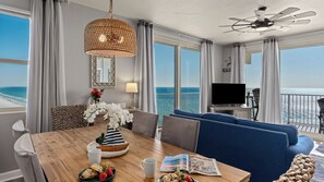 Una stanza con un tavolo da pranzo in legno, un divano blu e una TV. Dalle ampie finestre si vede l'oceano.