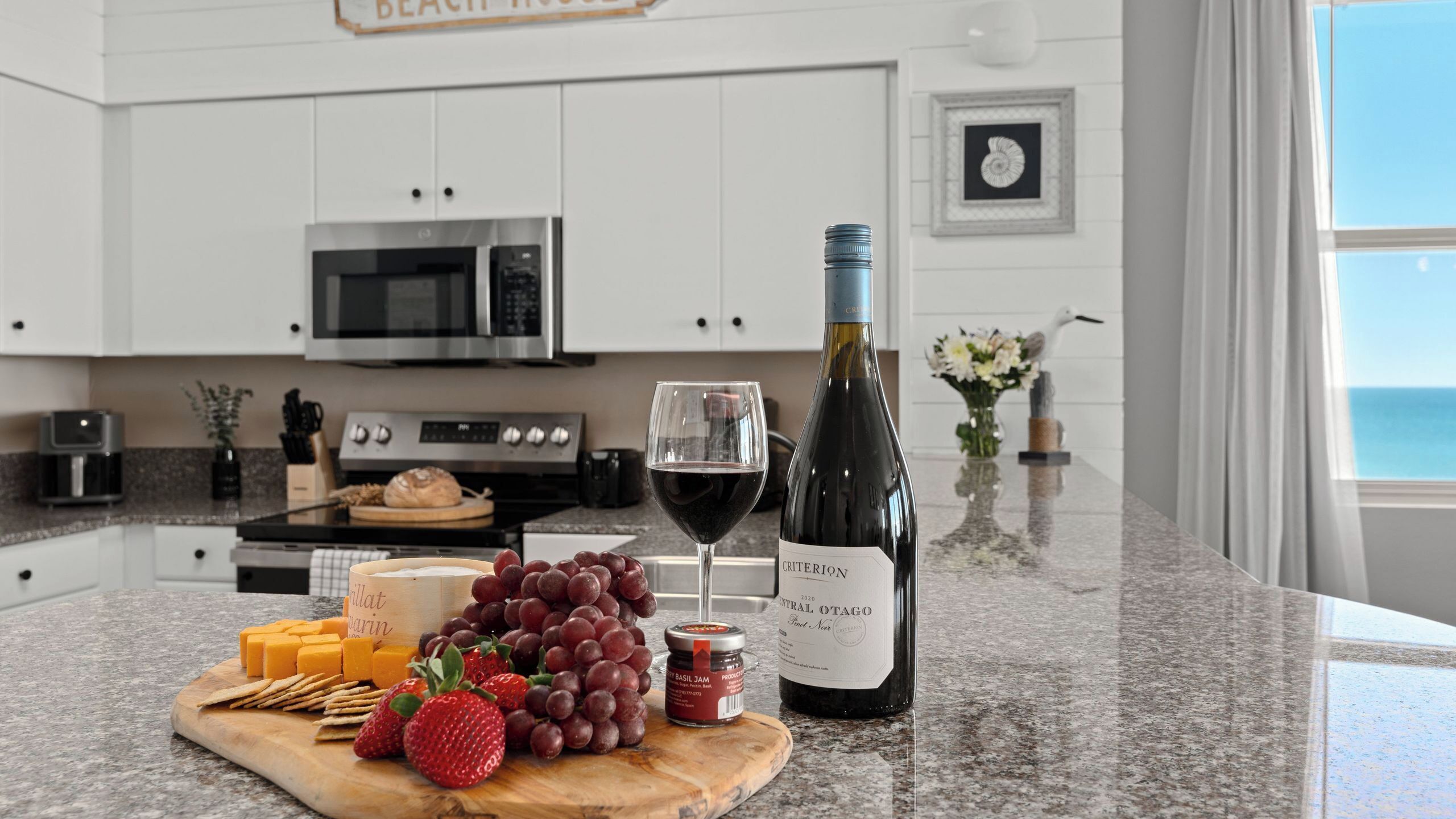 Una cocina con armarios blancos, un microondas, una estufa, una tabla de cortar con queso, uvas, fresas, una botella de vino y un frasco de mermelada.