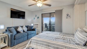 A bedroom with a bed, a blue sofa, a ceiling fan, a TV, and a balcony with a chair. - BayCation Walk to Jolly Roger + Beach (Ocean City)