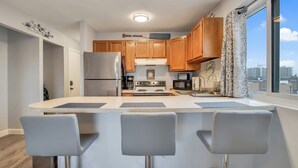A modern kitchen with wooden cabinets, a central island with bar stools, stainless steel appliances, and a large window offering a city view. - BayCation Walk to Jolly Roger + Beach (Ocean City)