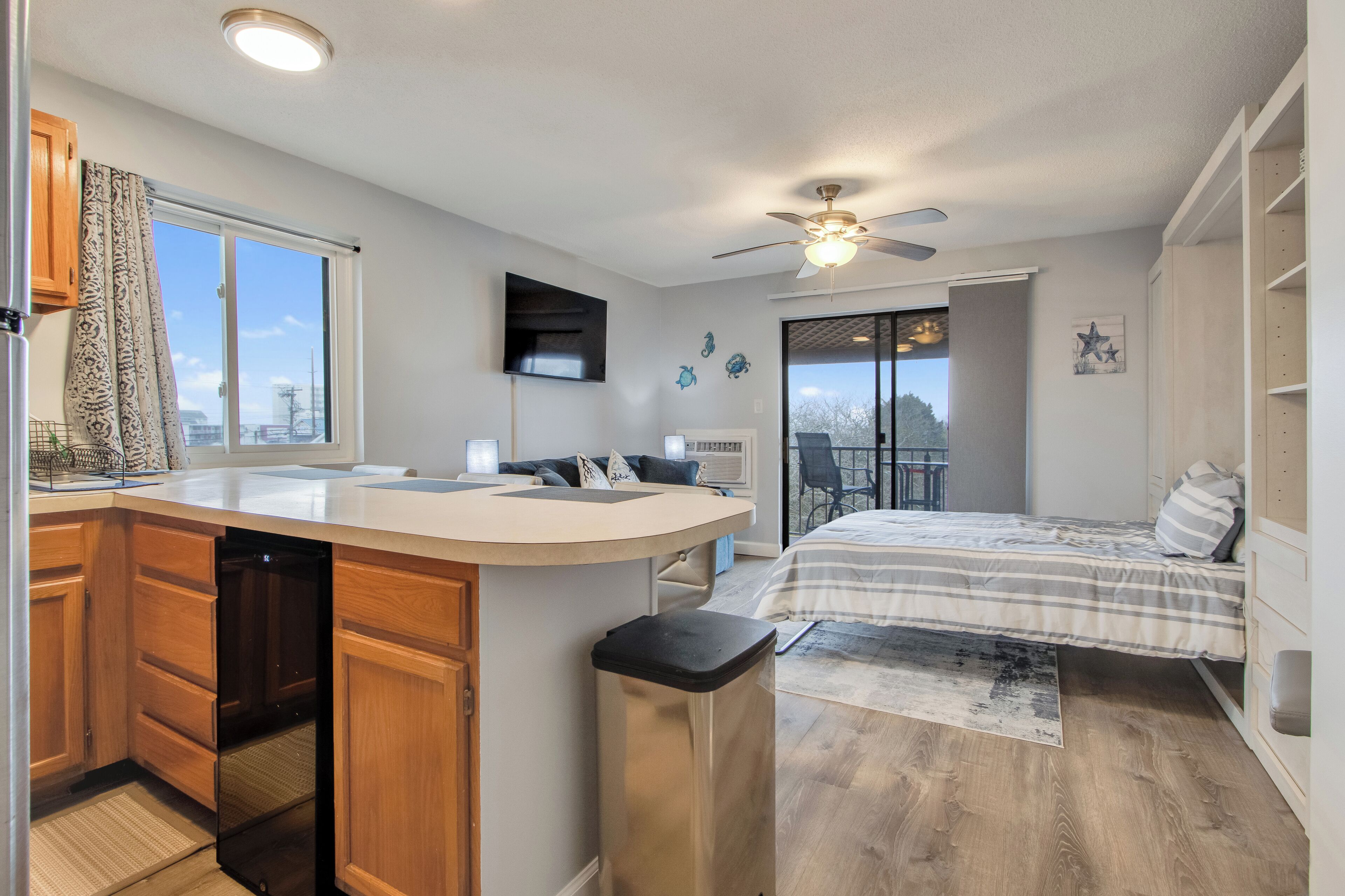 A compact apartment with a kitchenette, a bed, a ceiling fan, and a view of the outdoors.