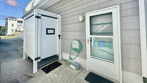 A building with a white door and window, a small outdoor area with a metal grate, and a green hose on the ground. - Gardens Single-Family Home • Ocean Views • Dog Considered • FRIDAY TO FRIDAY (Ocean City)