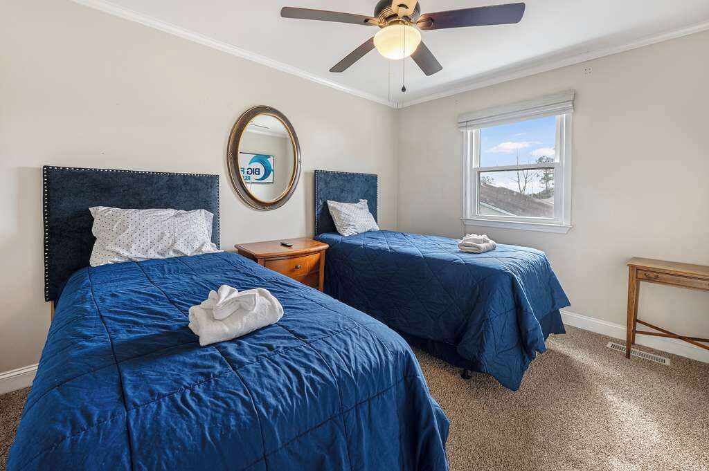 Habitación de hotel con dos camas, ventilador de techo, ventana con vistas y una mesita de noche de madera.
