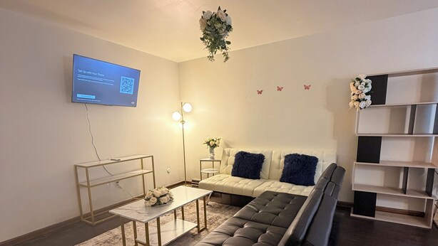En moderne stue med en sort sofa, et lille sofabord, et fjernsyn monteret på væggen og en gulvlampe.