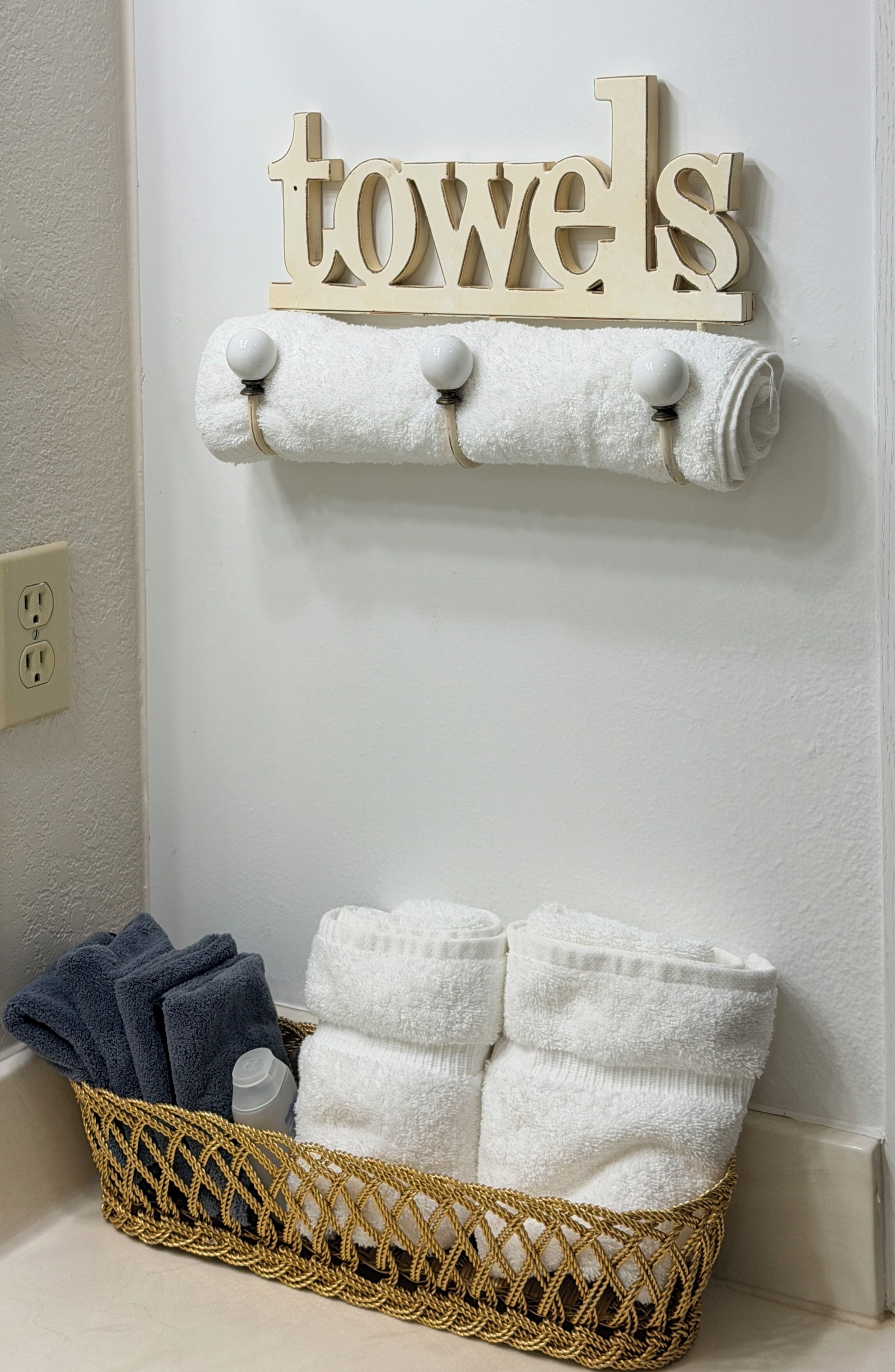 A bathroom with a towel rack holding three white towels, a wicker basket with additional towels, and a wall-mounted sign that reads "towels".
