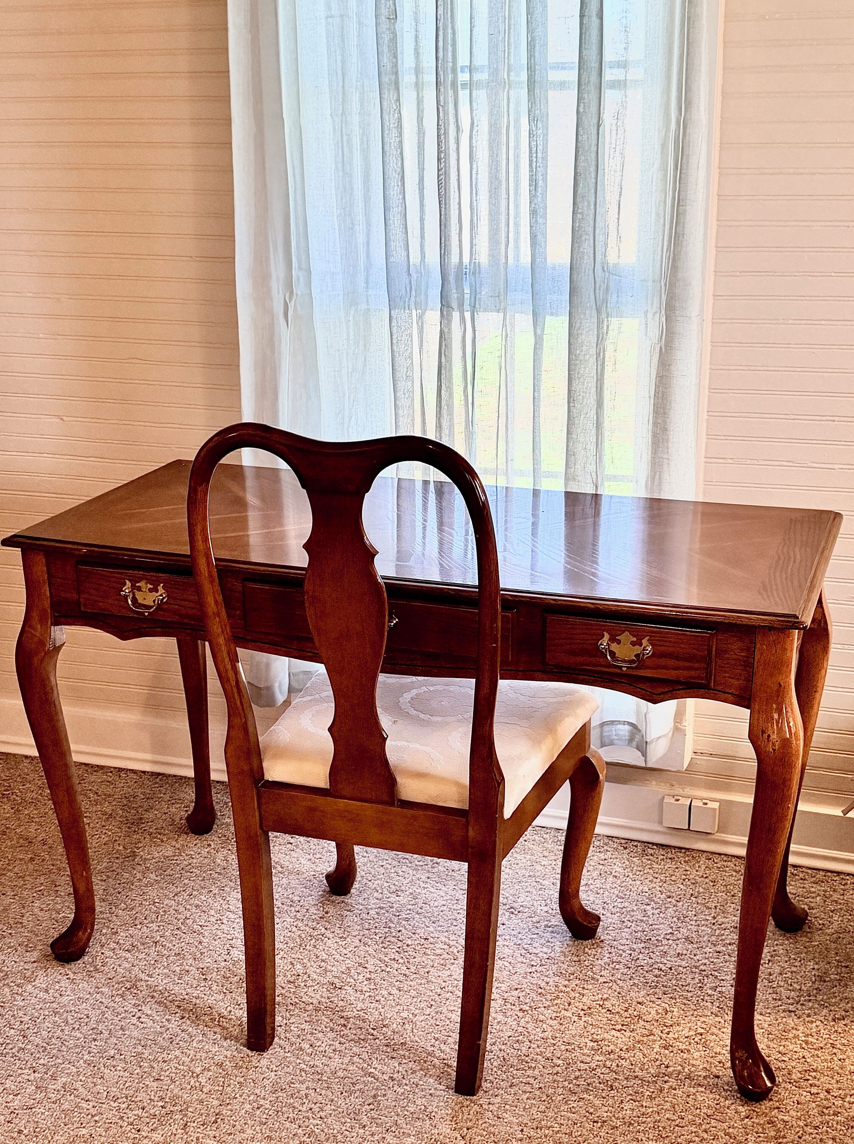 Una mesa de madera para comer con dos sillas, una ventana con cortinas ligeras y un suelo alfombrado.