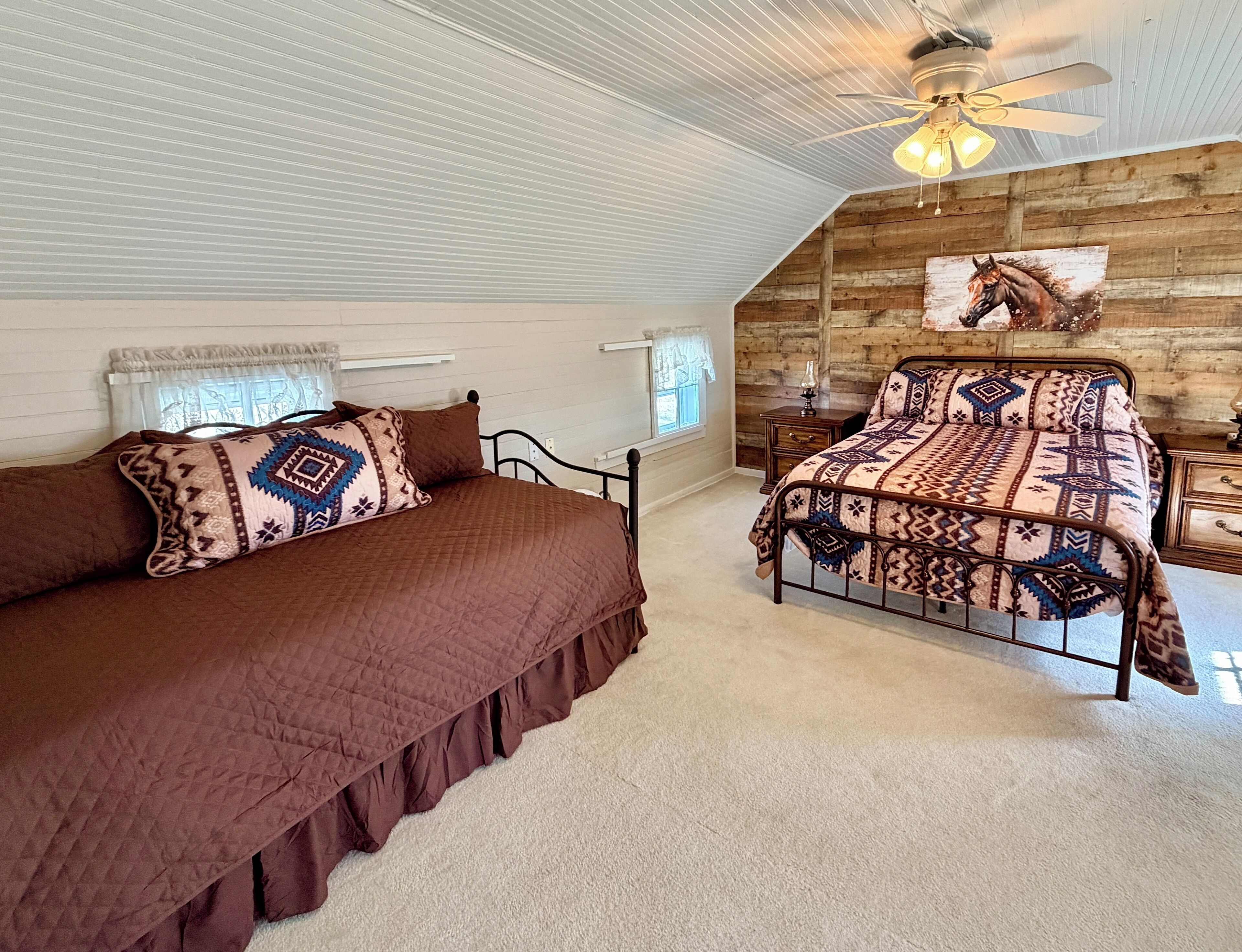 Een slaapkamer met een groot bed, een kleiner bed, een plafondventilator en een schilderij van een paard aan de muur.