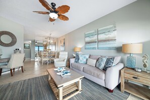 A living room with a ceiling fan, a sofa, a coffee table, a lamp, and artwork on the wall. - The Palms West 515 | Beachfront | Pools | My Beach Getaways (Orange Beach)