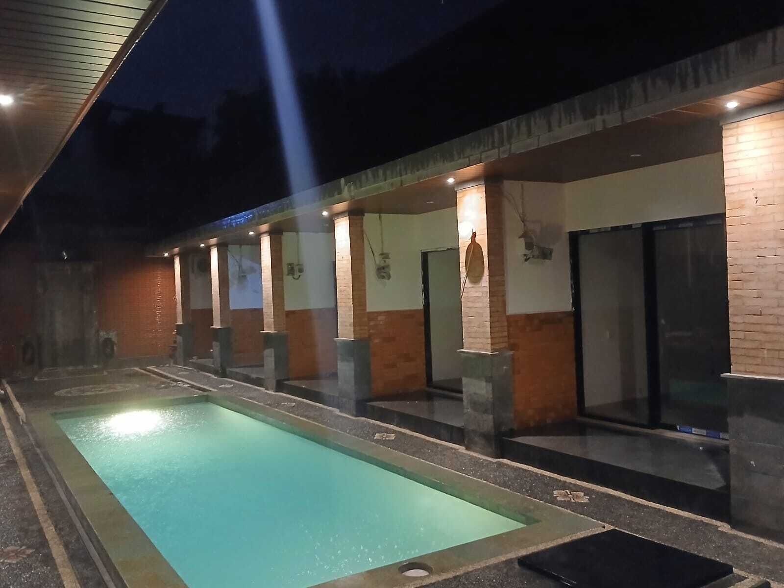 A swimming pool with a tiled deck, adjacent to a building with multiple rooms and a covered area.