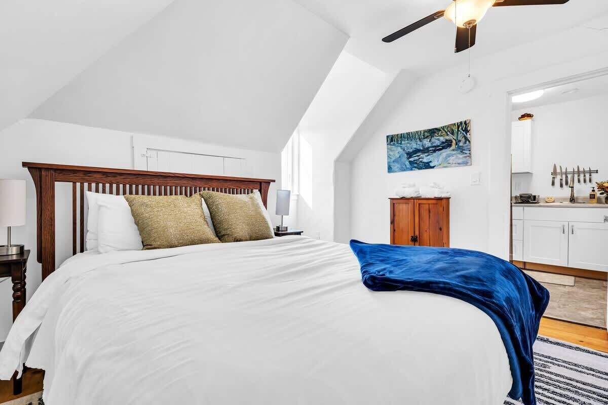 Een slaapkamer met een houten hoofdbord, witte beddengoed, een blauwe plaid en een schilderij van een boslandschap.
