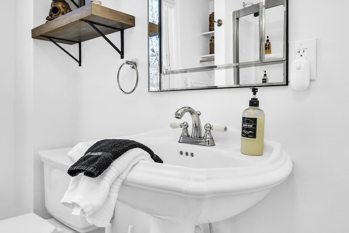 Une salle de bain avec un lavabo blanc, un miroir, une serviette, un distributeur de savon et une étagère contenant des articles de toilette.