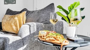 Una sala de estar moderna con un sofá gris, una mesita redonda con una tabla de quesos y copas de vino blanco, y una planta en maceta.