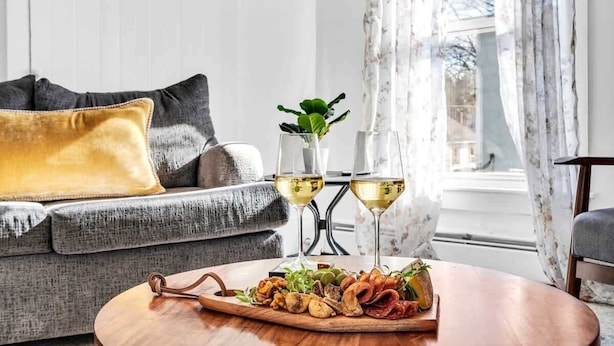 Una sala de estar acogedora con un sofá gris, una mesa de centro de madera con una tabla de embutidos y dos vasos de vino blanco sobre una mesita auxiliar.